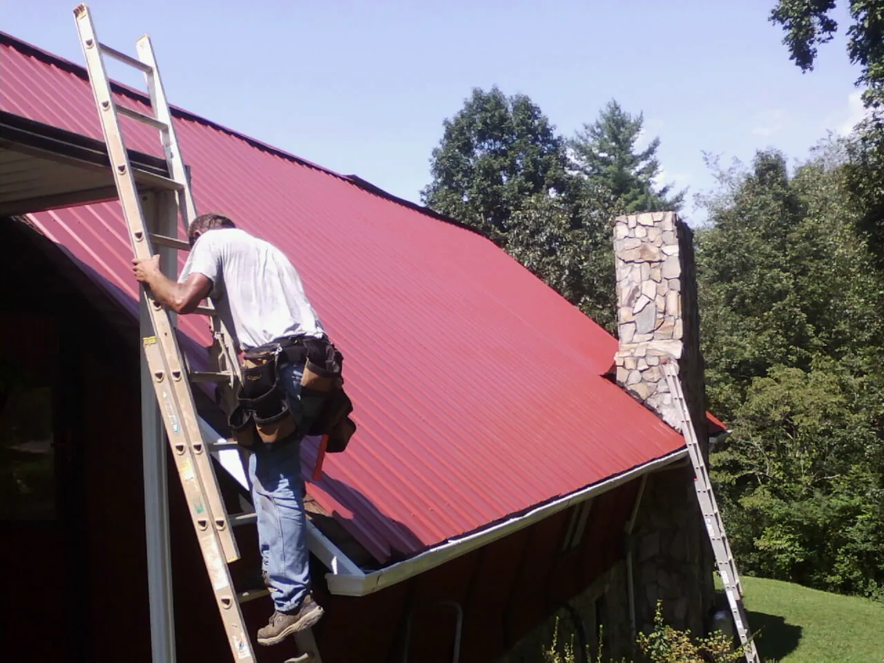 Roofer on a ladder beside a red metal roof and stone chimney — Soffit & Fascia Repair by Roofing Brothers in Piermont